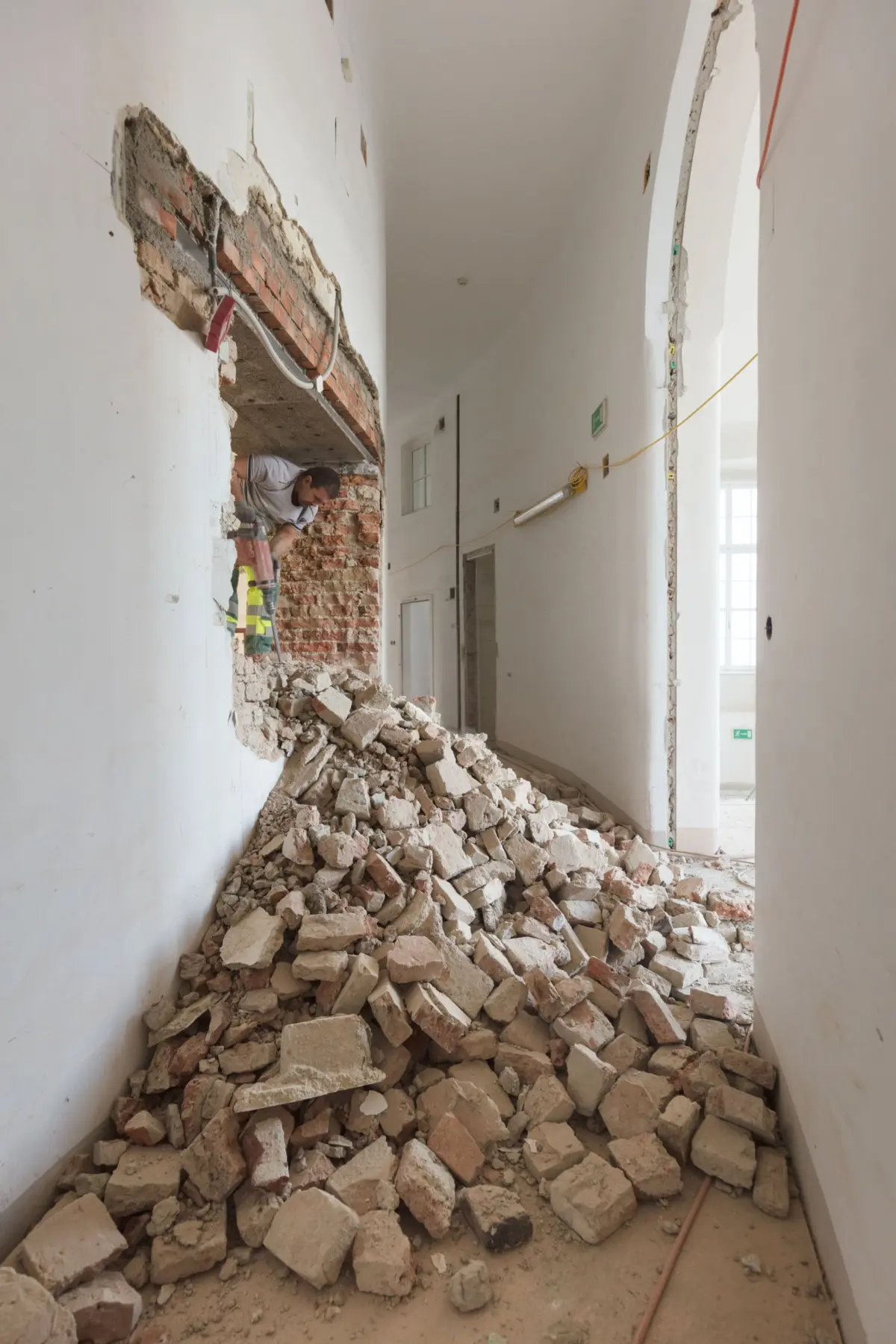 Construction worker uses a jackhammer to breach a brick wall in a long hallway, rubble scattered on the floor.