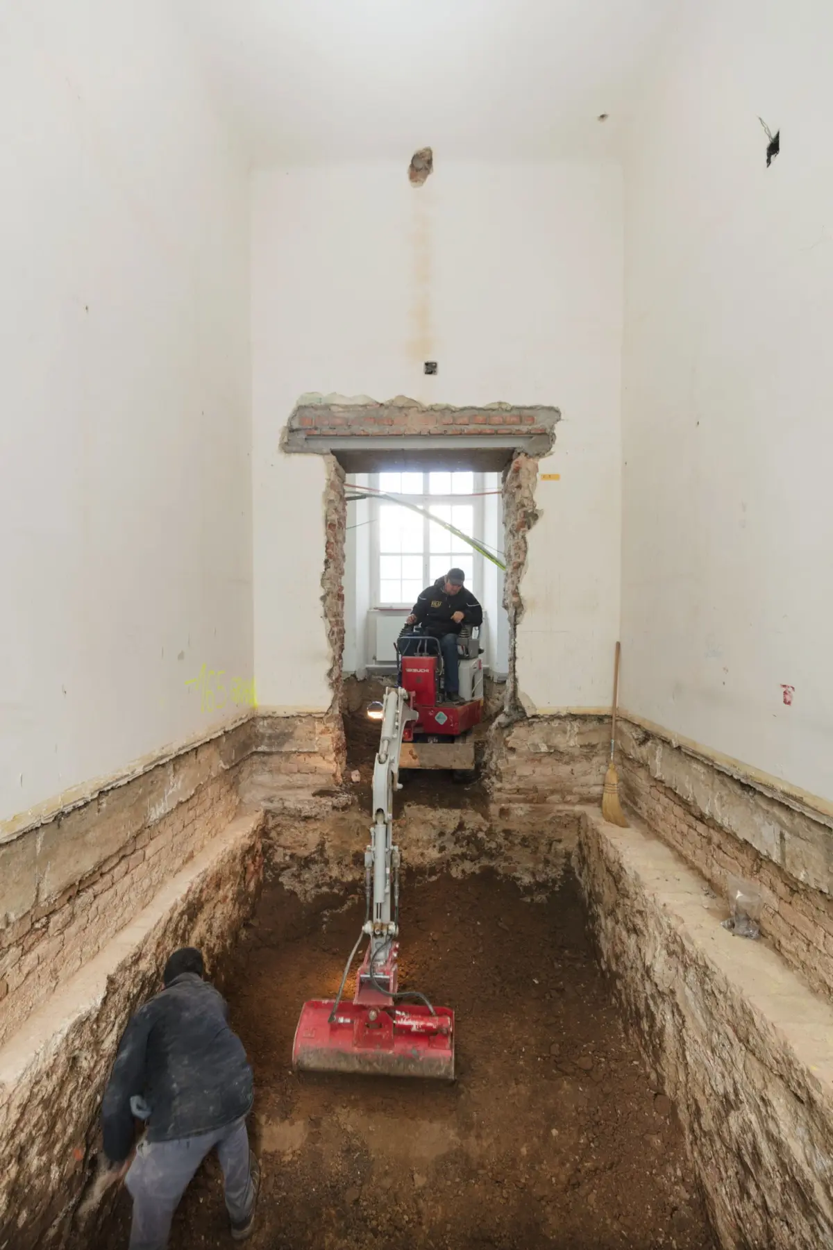 Two construction workers operate a small excavator inside a deep interior trench in a building under renovation; exposed brick doorway.