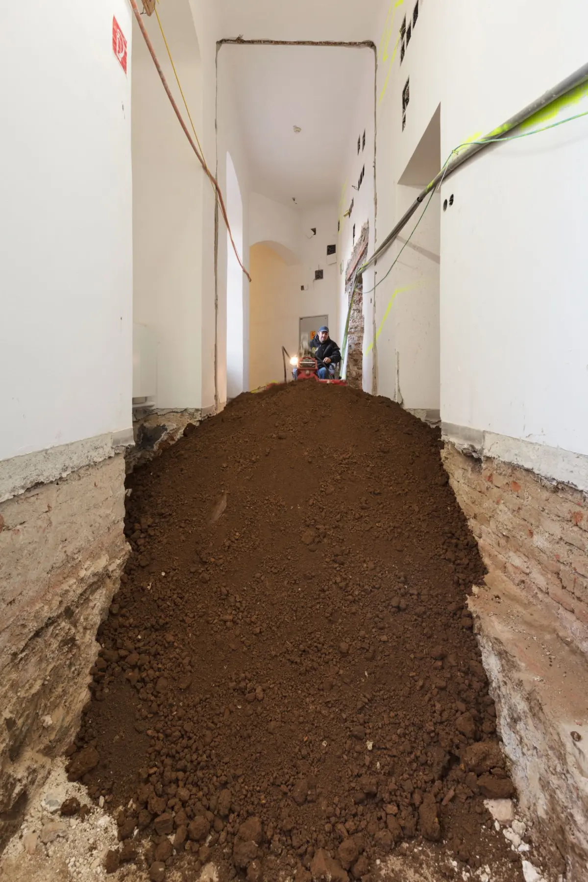 Construction worker operating excavation equipment in a narrow indoor hallway, digging a dirt trench.