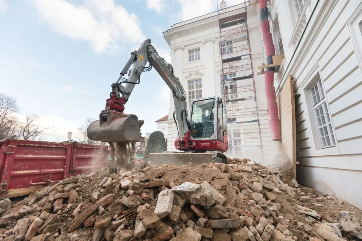 Excavator at a construction site removing rubble beside an ornate white building with scaffolding