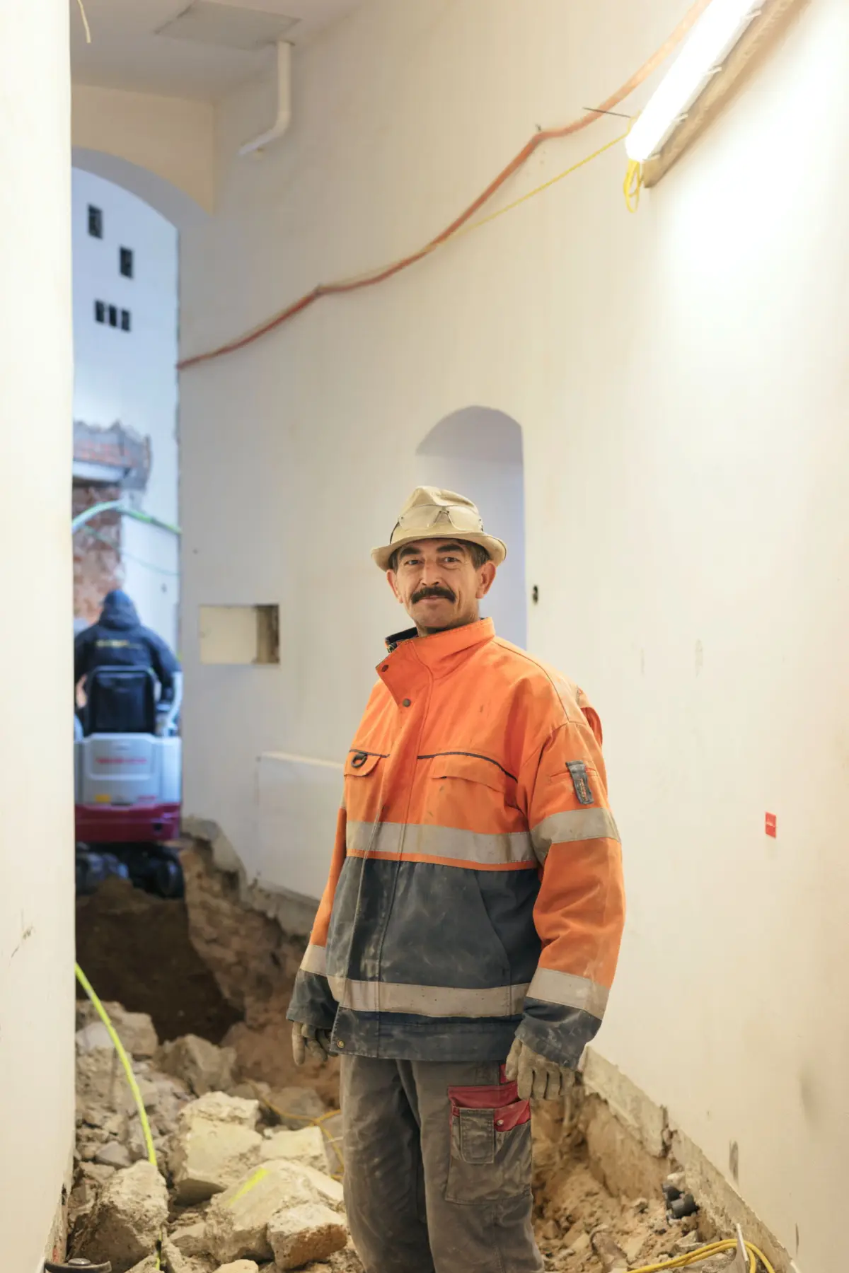 Construction worker in orange reflective jacket and beige hard hat stands in a hallway under construction, rubble on the floor.