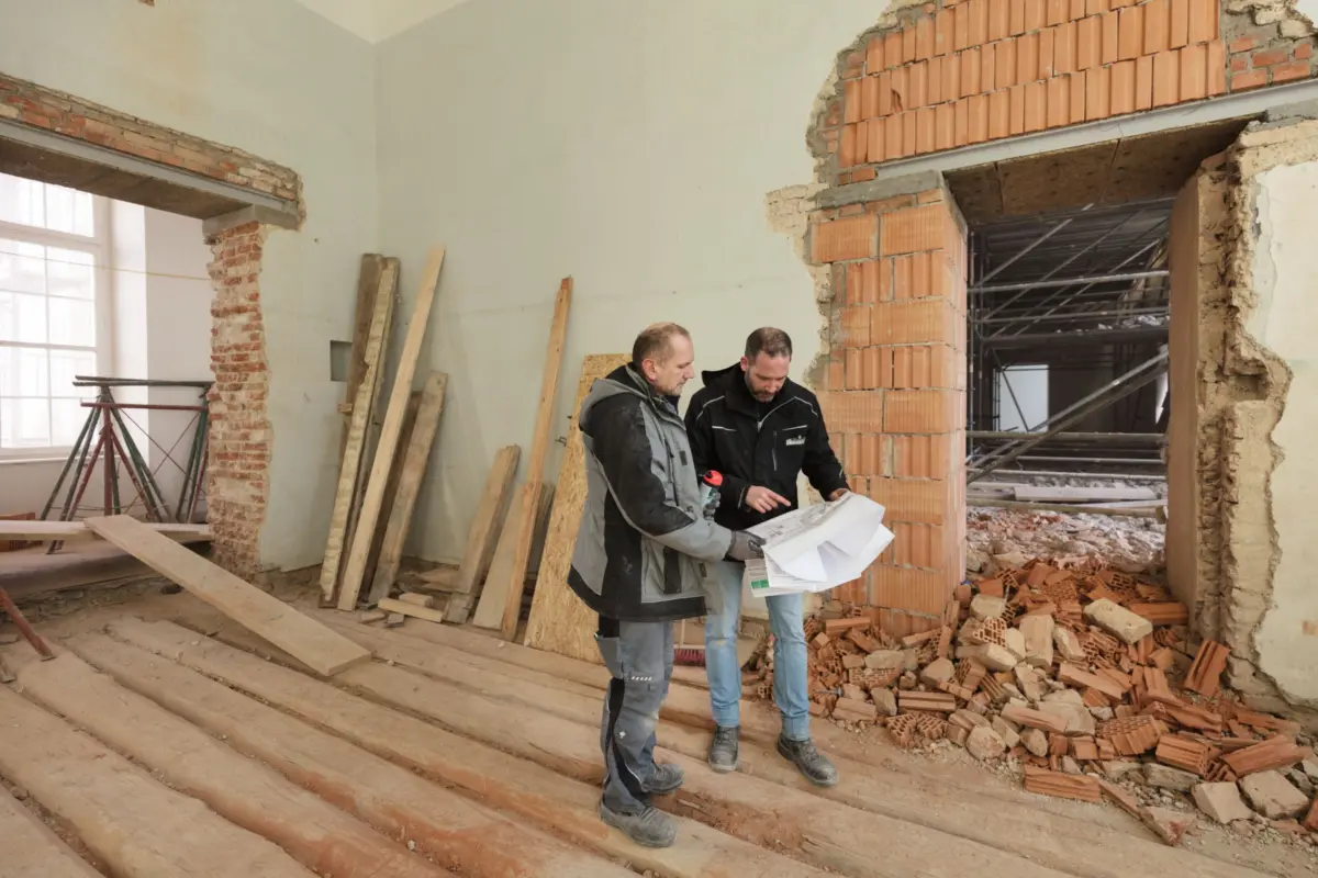 Two construction workers review blueprints inside a partially demolished room with exposed brick and rubble.