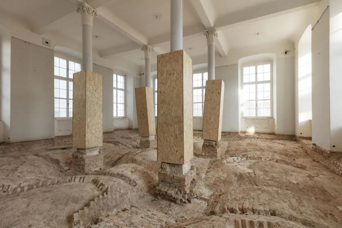 Historic interior under renovation: tall white columns with ornate capitals, upright OSB panels around them, and exposed brick floor near large windows.
