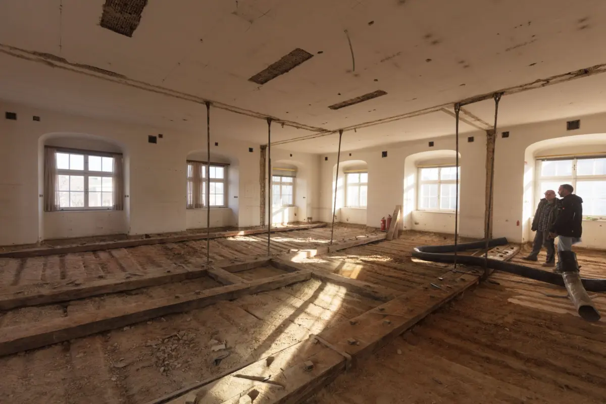 Two construction workers stand in a sunlit, half-demolished hall with exposed trenches, vertical supports, hoses, and arched windows.