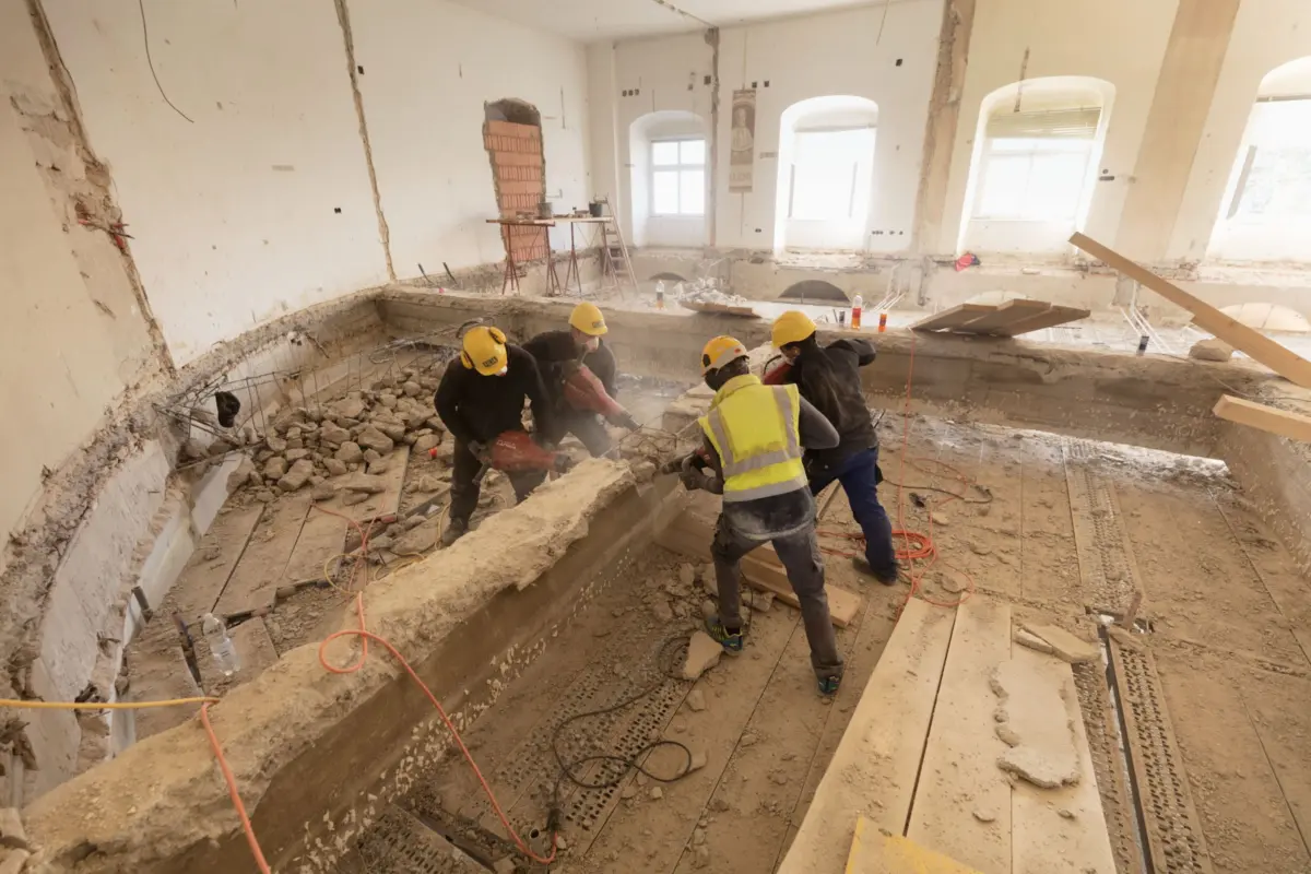 Construction crew in yellow hard hats jackhammering rubble inside a partially demolished, windowed renovation site.