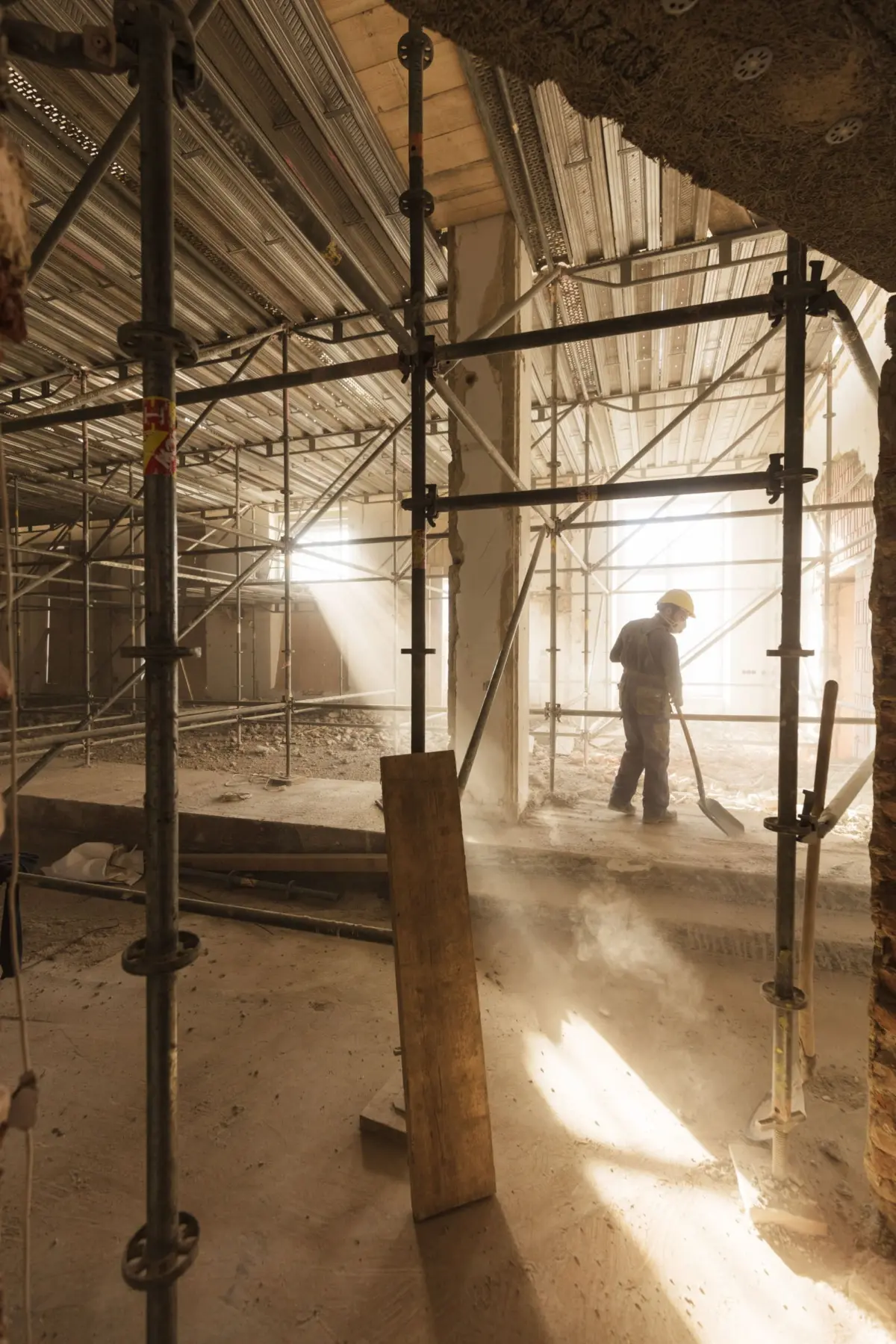 Interior construction site with metal scaffolding; a worker in a yellow hard hat and dust mask stands amid dust and debris.