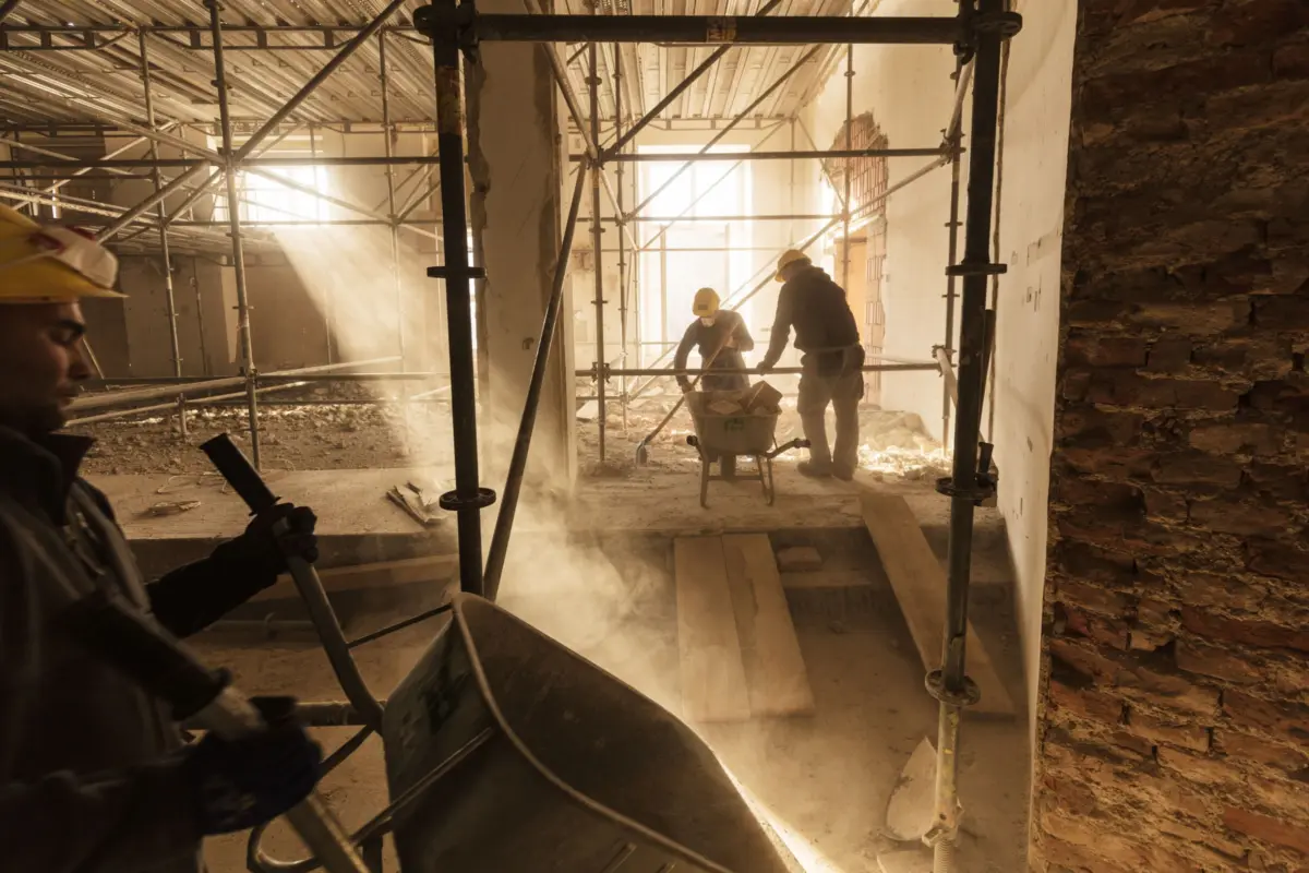 Two construction workers wearing yellow hard hats move debris beside a wheelbarrow on a dusty site with metal scaffolding.