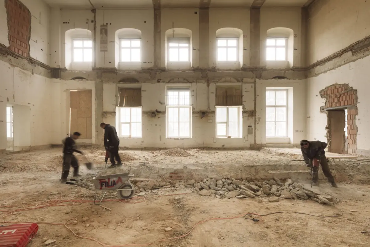 Construction workers clearing rubble inside a derelict building with tall arched windows; wheelbarrow labeled FILA in foreground.
