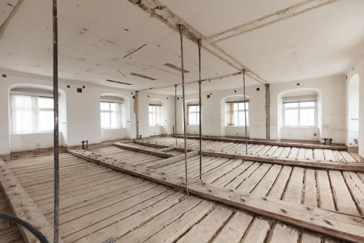 Spacious unfinished room under renovation with exposed ceiling patches, black metal support poles, bare wooden floor, and arched windows.