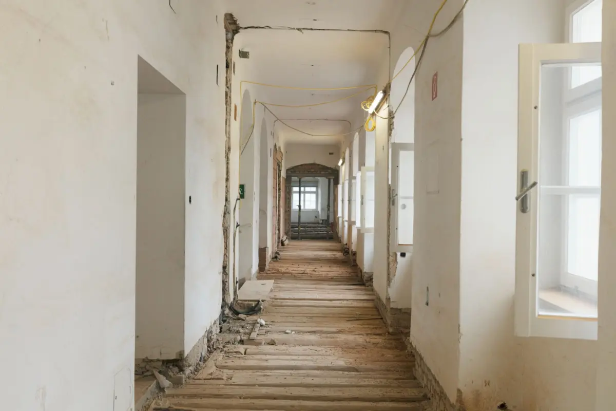 Long renovation hallway with exposed brick, hanging yellow cables, and debris on a wooden floor.