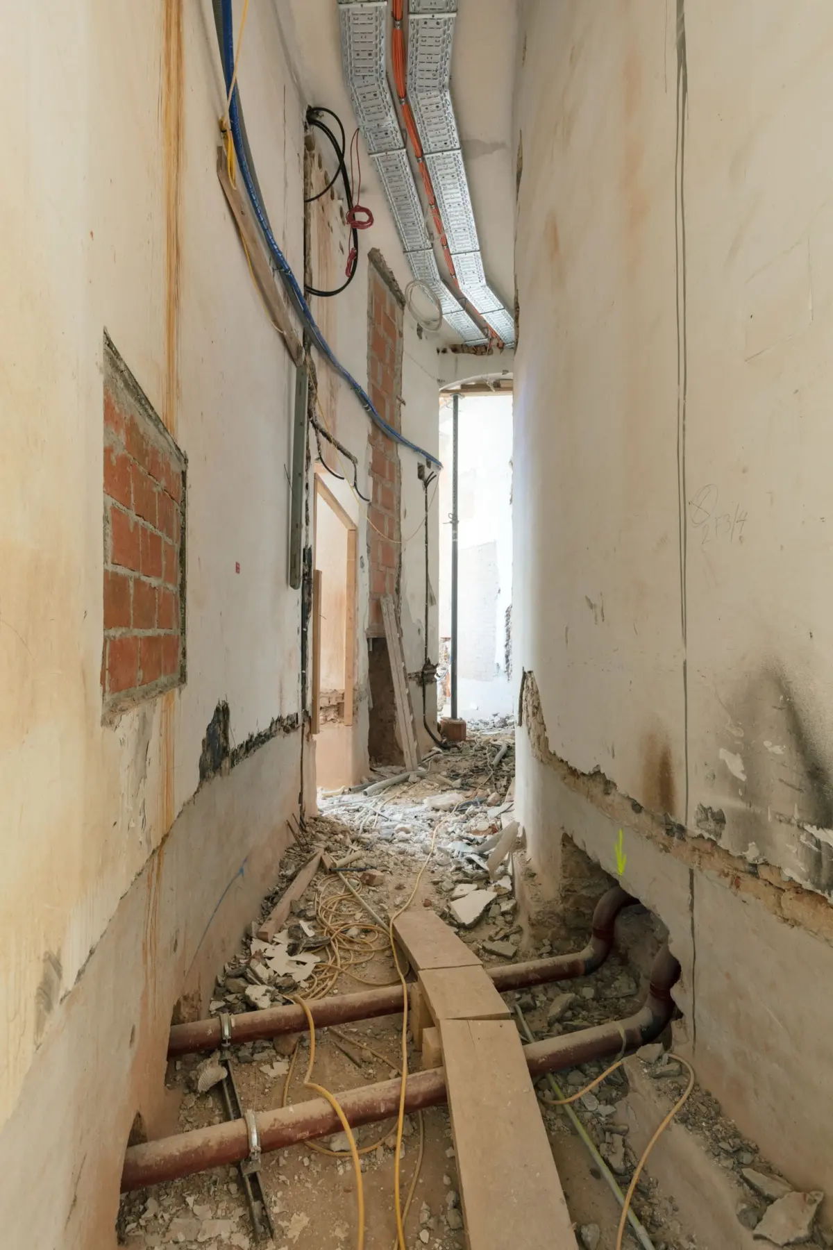 Construction hallway with exposed brick walls, overhead metal cable trays, tangled wires, and red pipes along the floor amid construction debris.