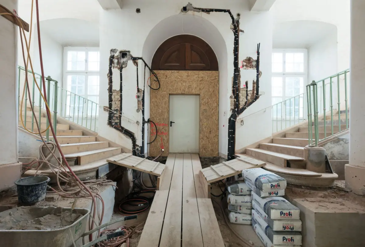 Renovation site inside a building: central arched doorway boarded with plywood, two staircases with green rails, exposed wires, cement bags.