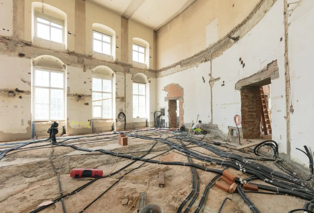 Construction worker crouches among tangled cables in a dilapidated brick-walled renovation hall with tall arched windows.