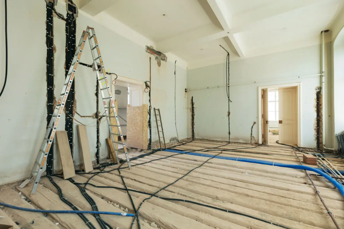 Interior renovation scene with exposed wiring and plumbing, ladders leaning against walls, and torn plaster in a partially demolished room.