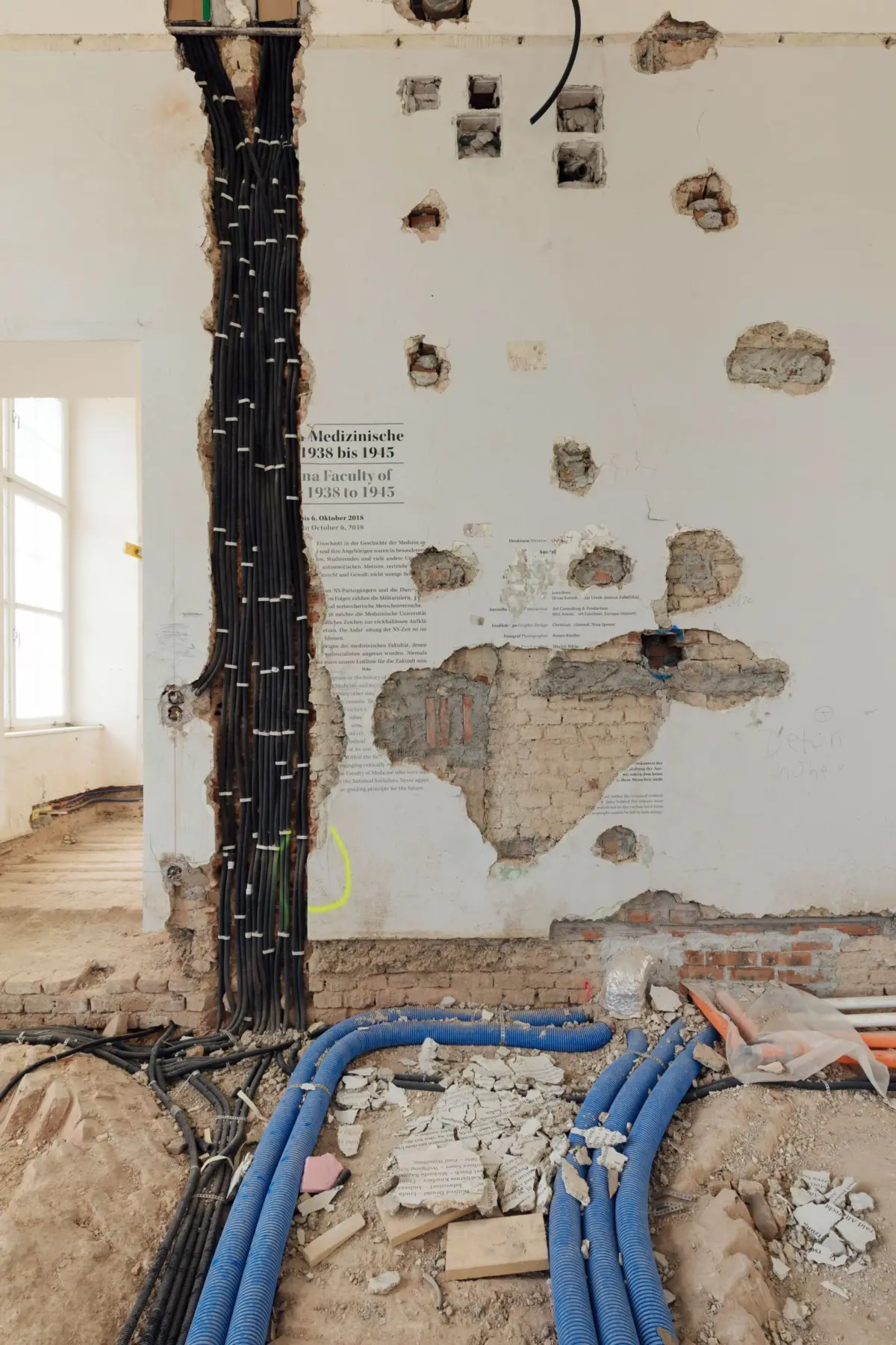 Interior of a building under renovation with exposed wiring: black cable bundles along a damaged wall and blue conduits on rubble.