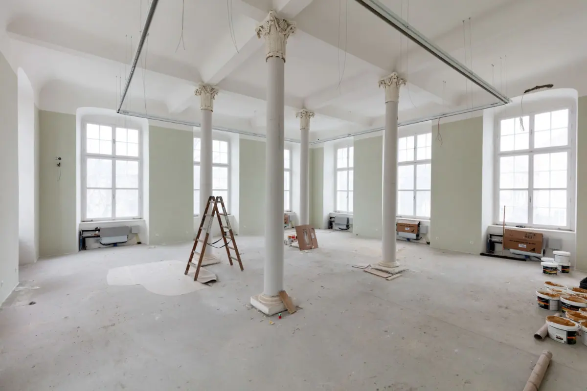 Interior renovation of a large room with tall Corinthian columns, large windows, ladders, and paint cans.