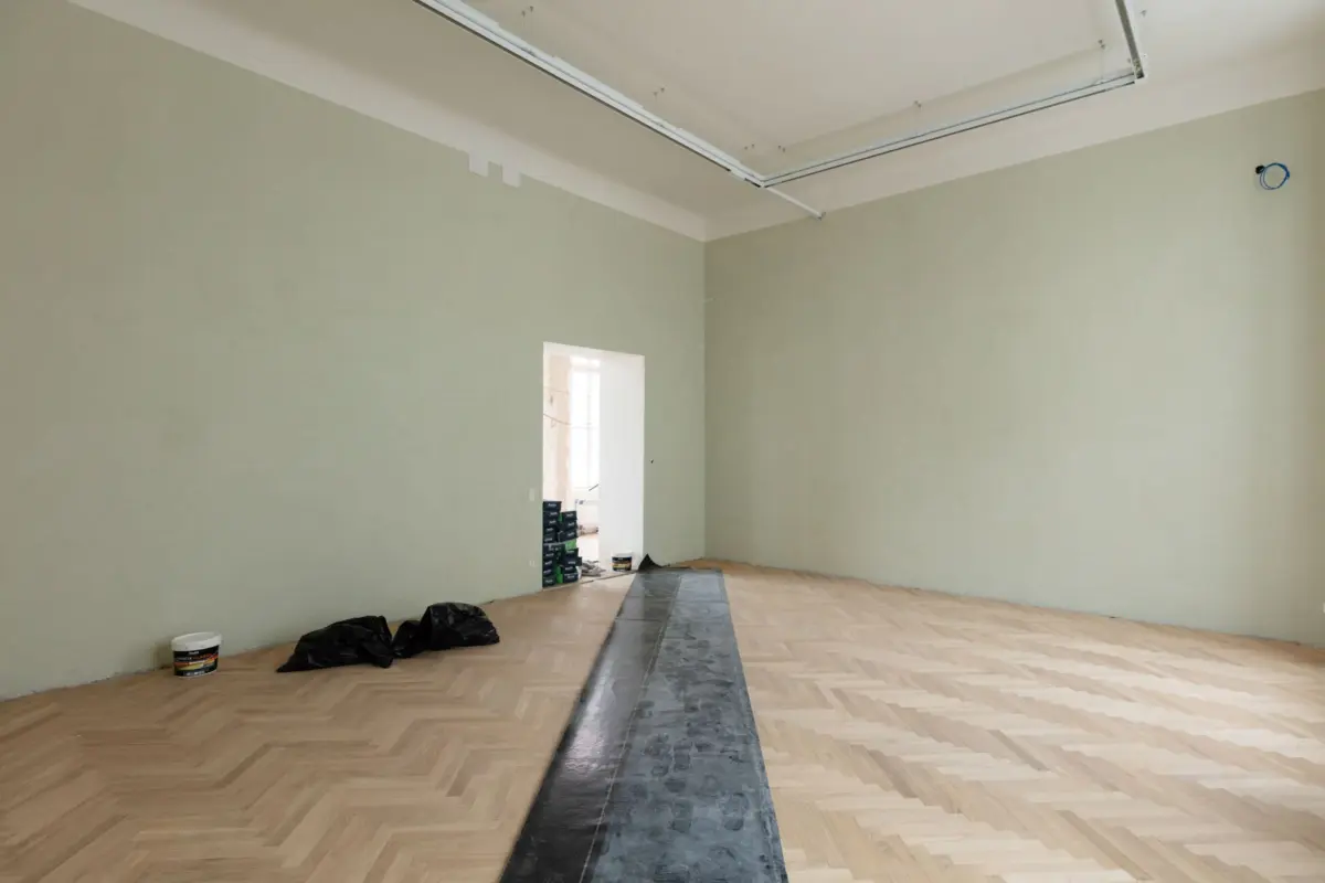 Spacious renovation room with pale green walls, herringbone parquet floor, exposed ceiling pipes, and paint cans by the doorway.