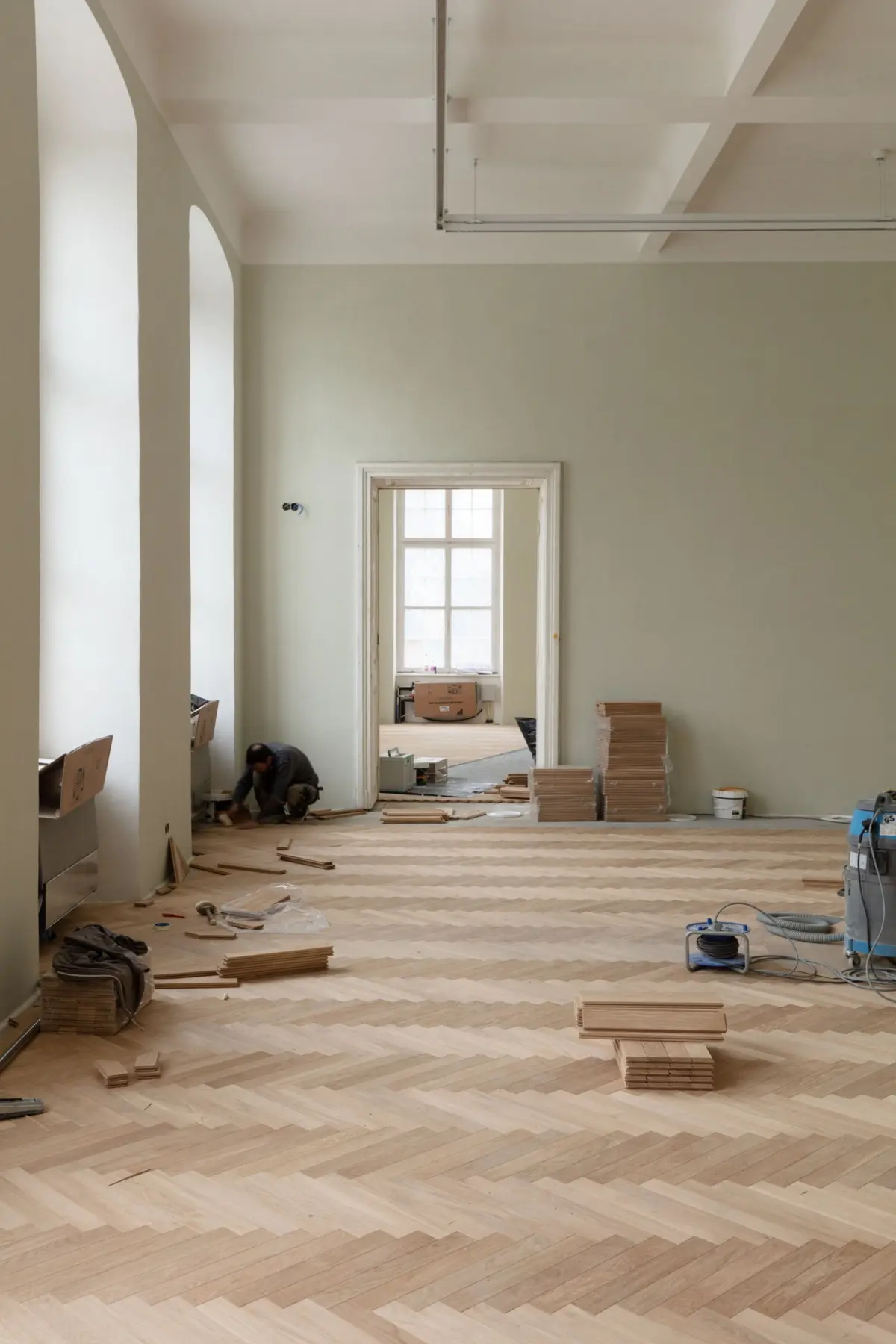 Construction worker kneels to lay herringbone wooden flooring in a pale-green room with arches and a doorway.