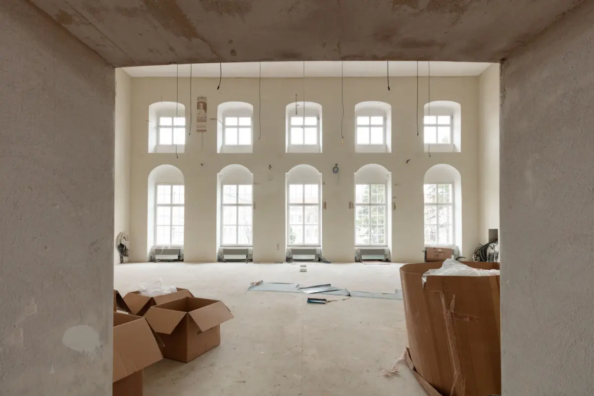Large unfinished hall with arched windows and exposed wiring; construction debris and cardboard boxes scattered.
