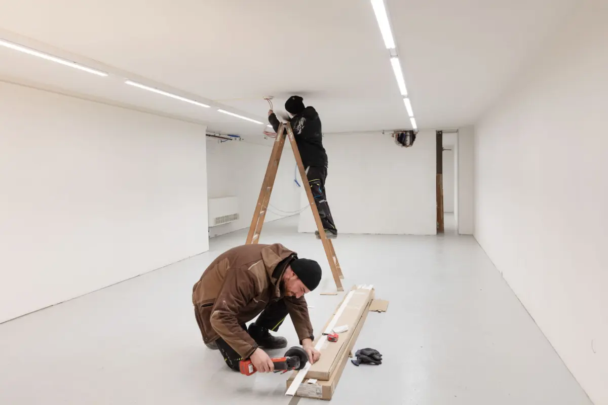 Two construction workers in a white, empty room: one on an A-frame ladder wiring the ceiling, the other kneels cutting wooden boards.