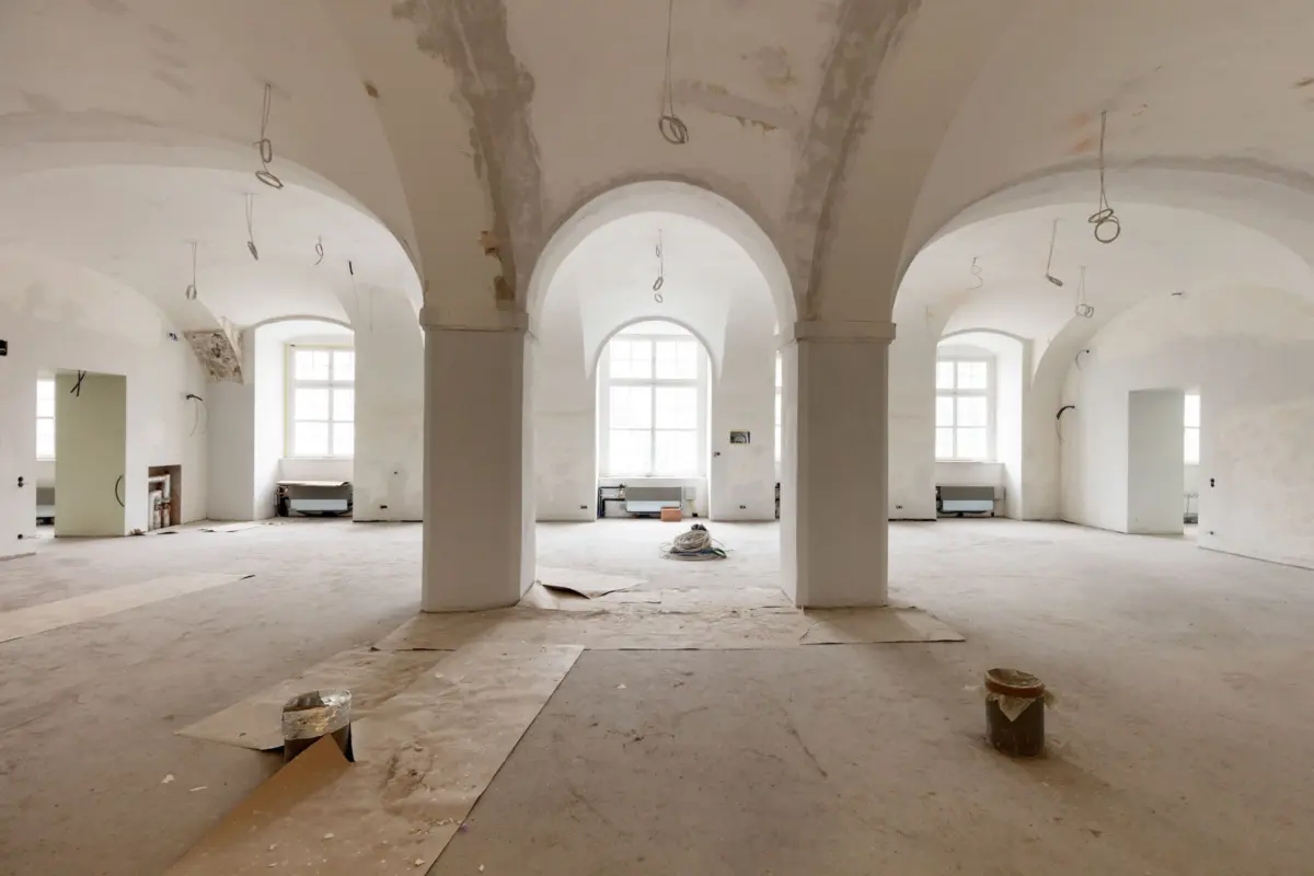 Interior of a large vaulted hall under renovation; arched ceilings, thick columns, hanging electrical wires, construction debris on the floor.