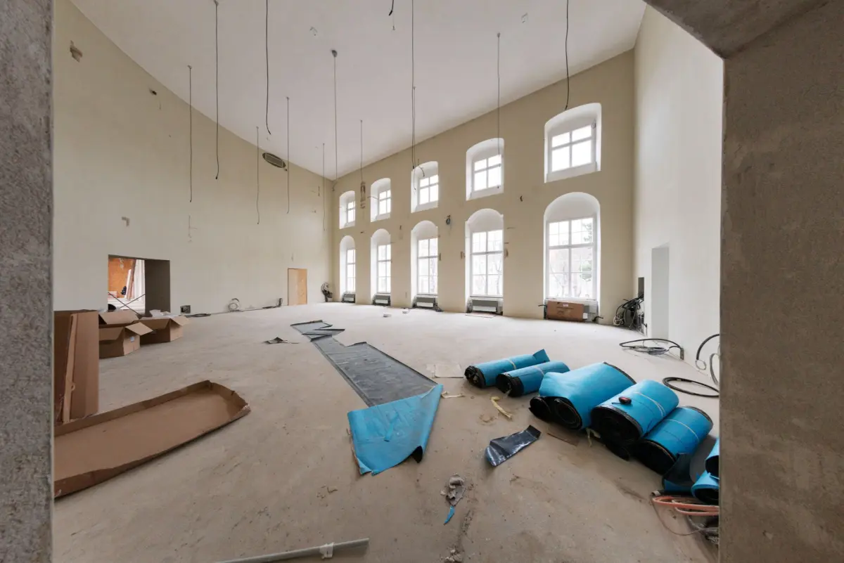 Under-construction hall with tall arched windows; hanging wires from ceiling, scattered cardboard and blue insulation rolls on the concrete floor.