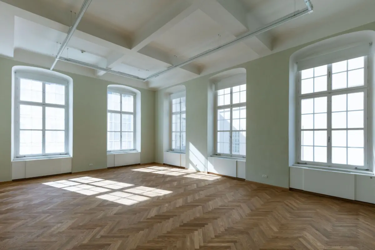Sunlit empty room with pale green walls, tall white-framed windows, and herringbone oak floor; exposed white ceiling beams.
