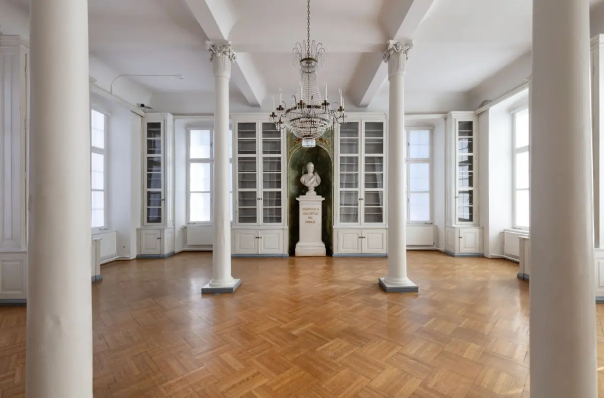 White neoclassical gallery with tall columns, marble bust on pedestal in green niche, glass cabinets, crystal chandelier, parquet floor.