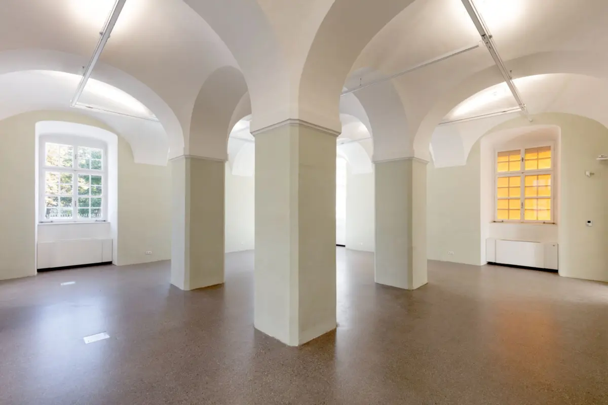 Empty hall with white vaulted arches and square columns; large windows along pale walls.