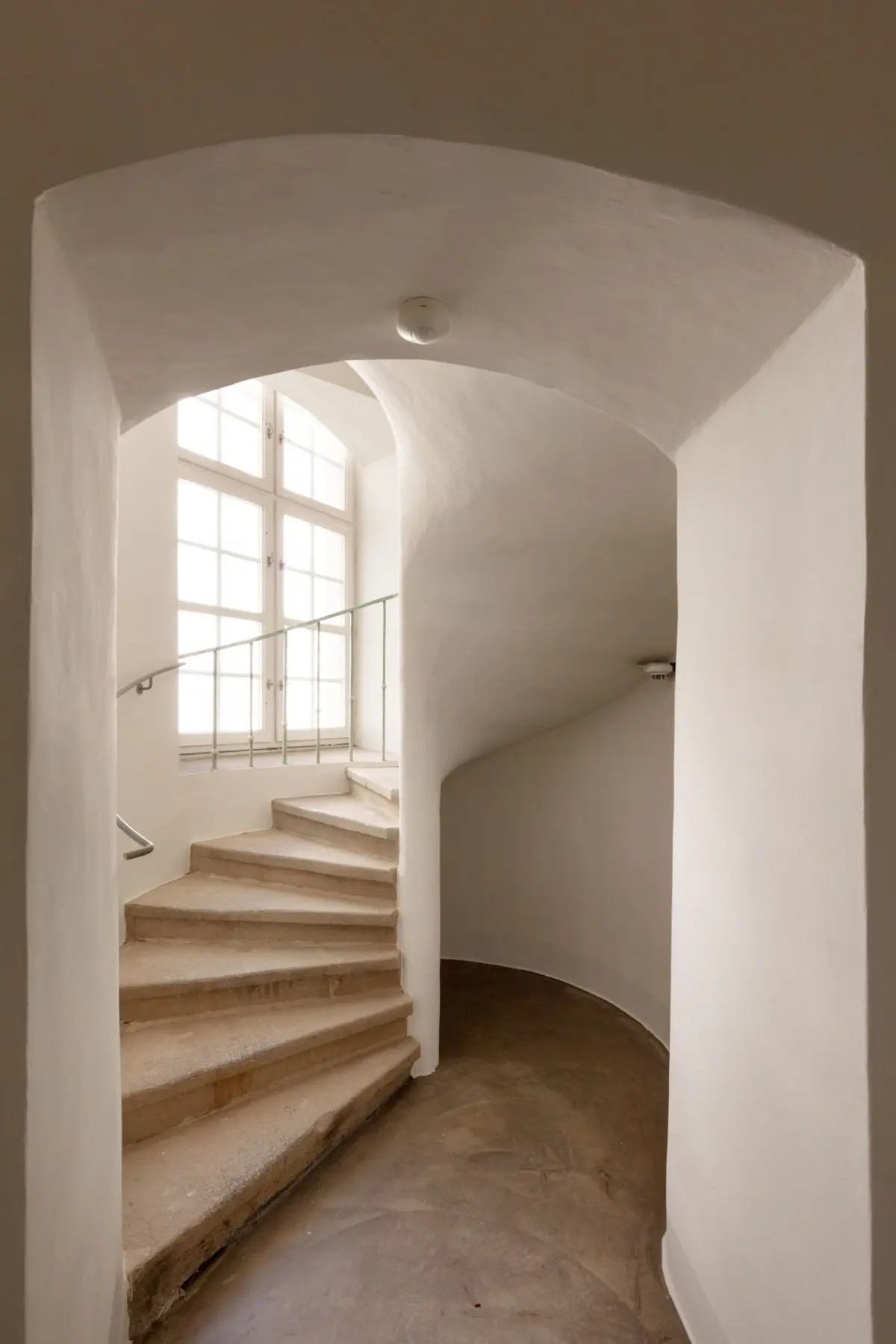 Curved white plaster staircase in a sunlit stairwell with an arched ceiling and large window at the landing.