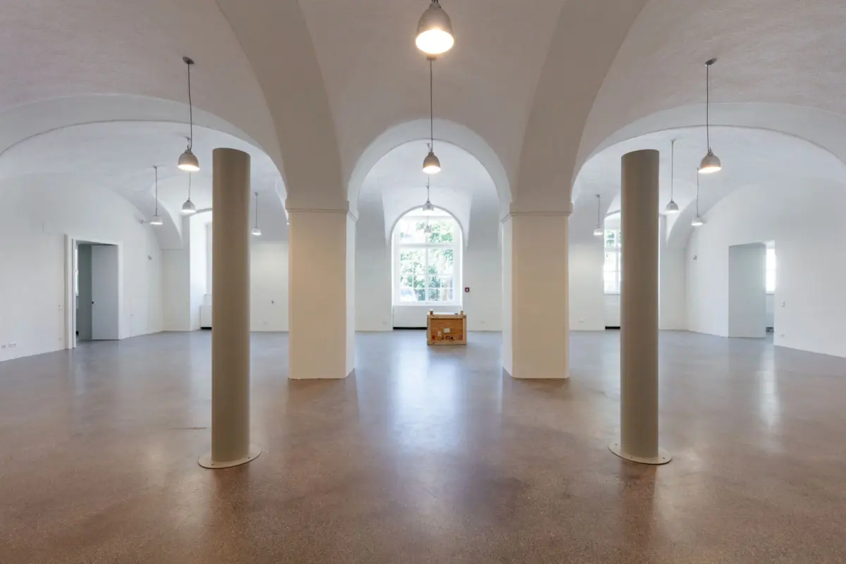 Empty spacious white gallery interior with cylindrical columns, vaulted arches, and hanging pendant lights; wooden crate at center.