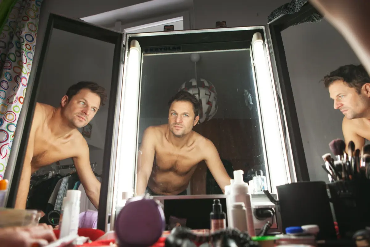 Three shirtless men lean toward a lit vanity mirror in a bathroom, surrounded by cosmetics and a colorful shower curtain.