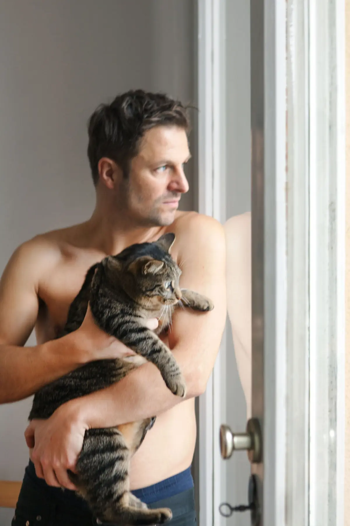Shirtless person holding a tabby cat by an interior window.