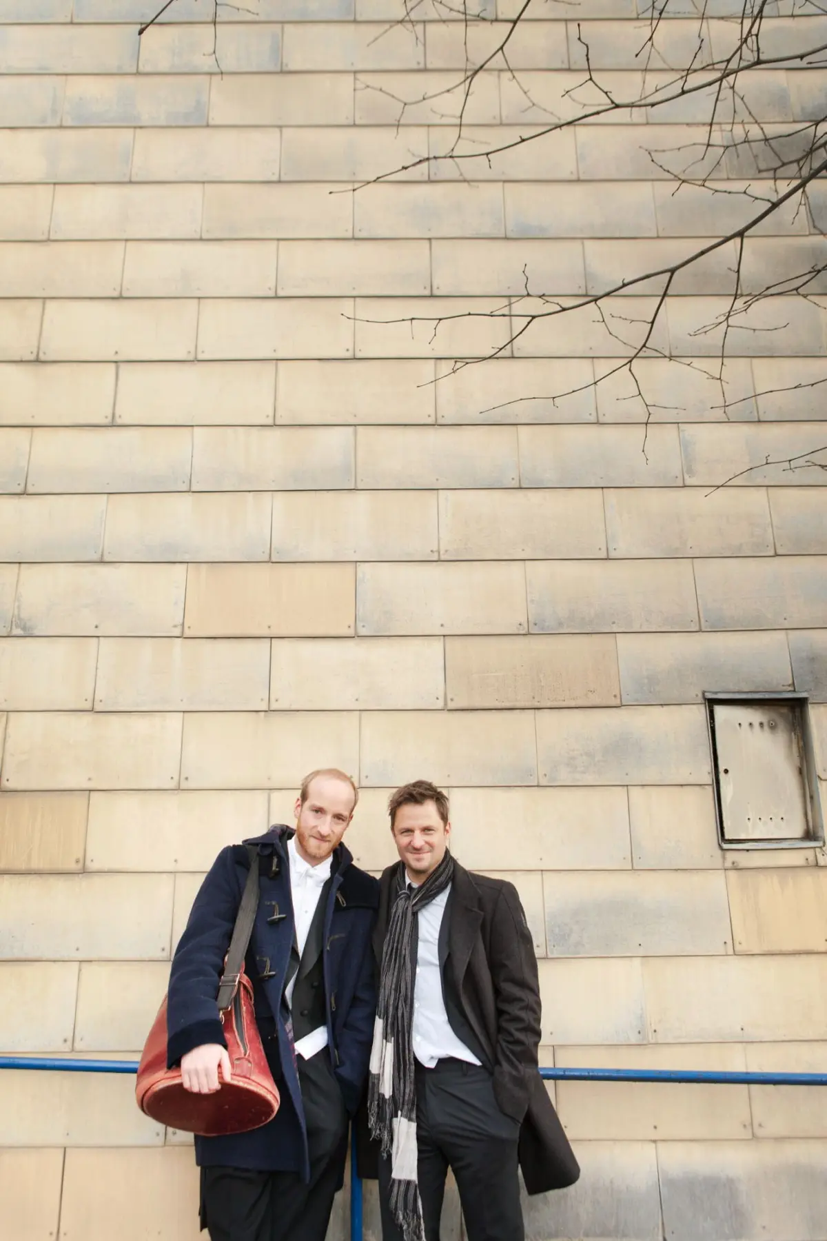 Two men pose together against a large beige tiled wall with bare tree branches overhead.