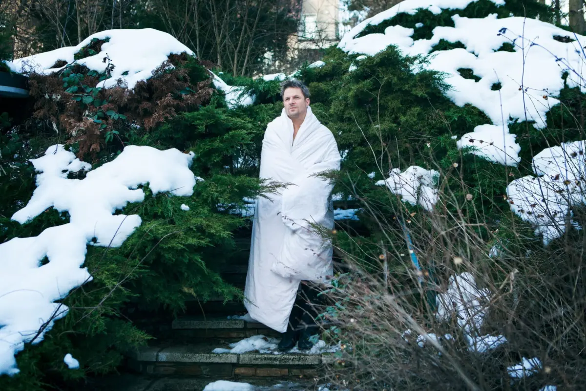Person wrapped in a white blanket stands among snow-covered evergreen shrubs in a garden.