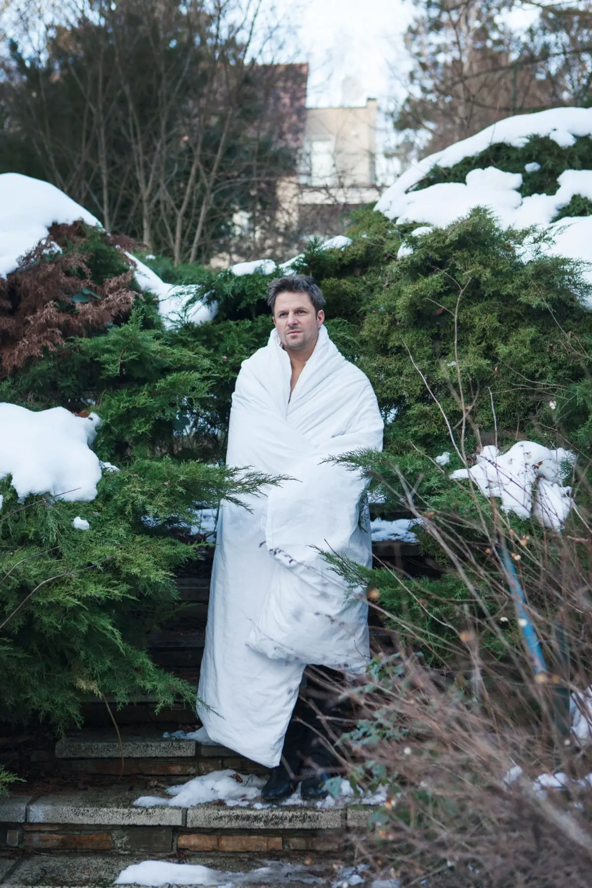 Person wrapped in a white blanket stands on stone steps among snow-covered evergreen shrubs outdoors.