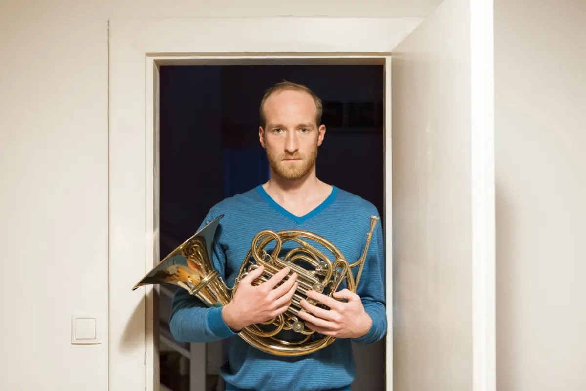Person in a blue striped sweater stands in a doorway, holding a French horn.