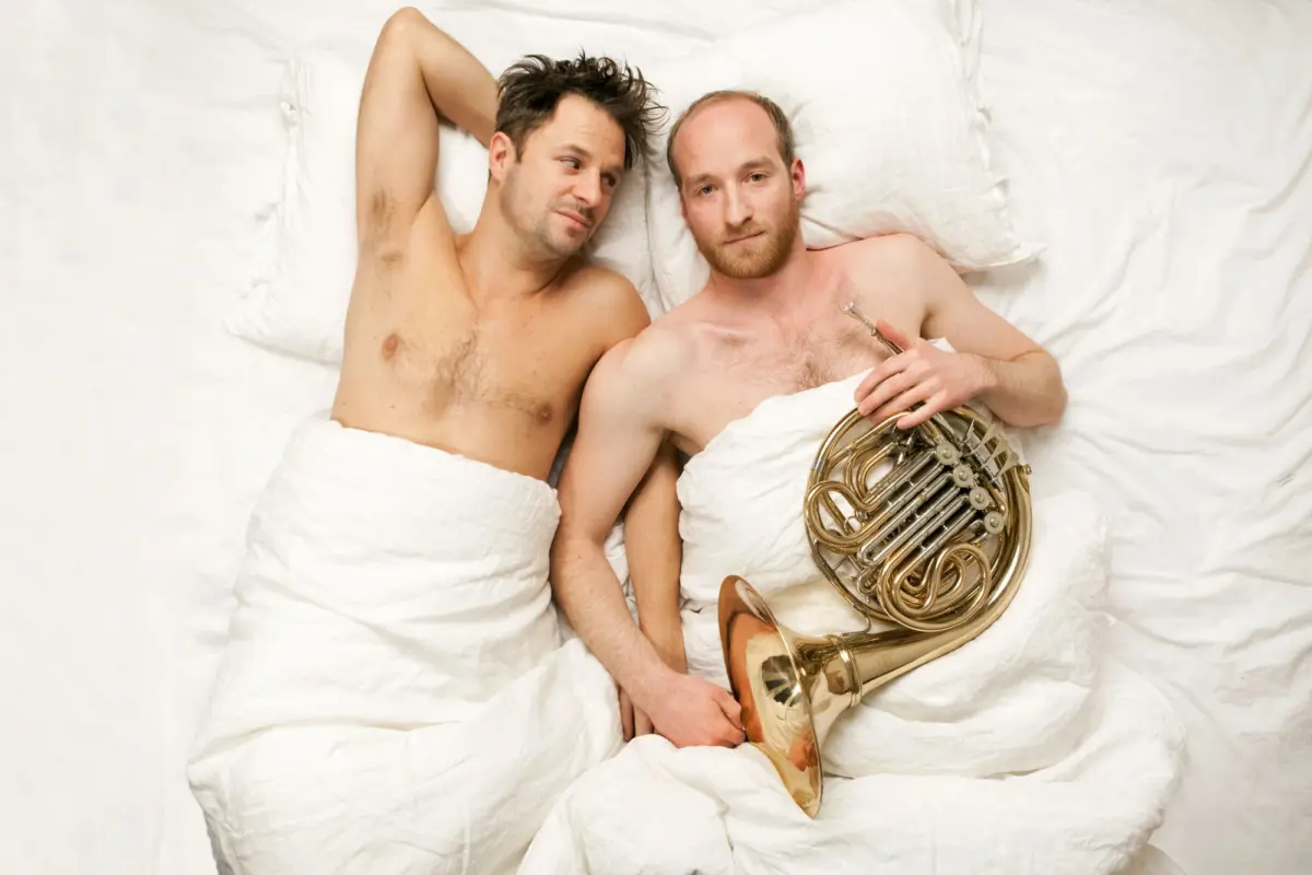 Two adults lying topless in a white bed; one rests with arm behind head while the other holds a French horn.