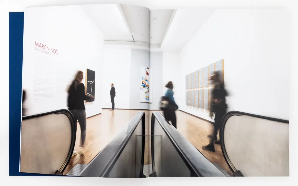 Escalator through a bright white art gallery; blurred visitors view wall-mounted abstract panels; signage reads 'MARTIN NOËL'.