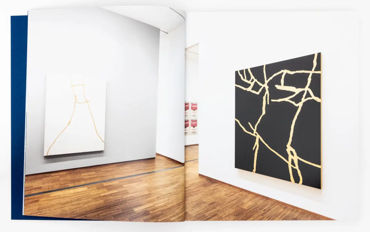 Open art book displaying a gallery scene: white walls with abstract canvases (white with gold lines, black with gold veins) on a wooden floor.
