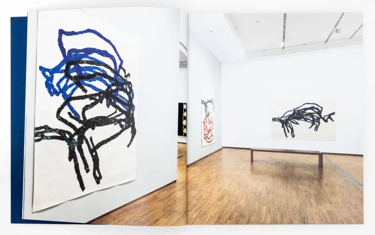 Open art book spread showing a blue and black abstract drawing, in a bright white gallery with wooden floor and wall-mounted artworks.