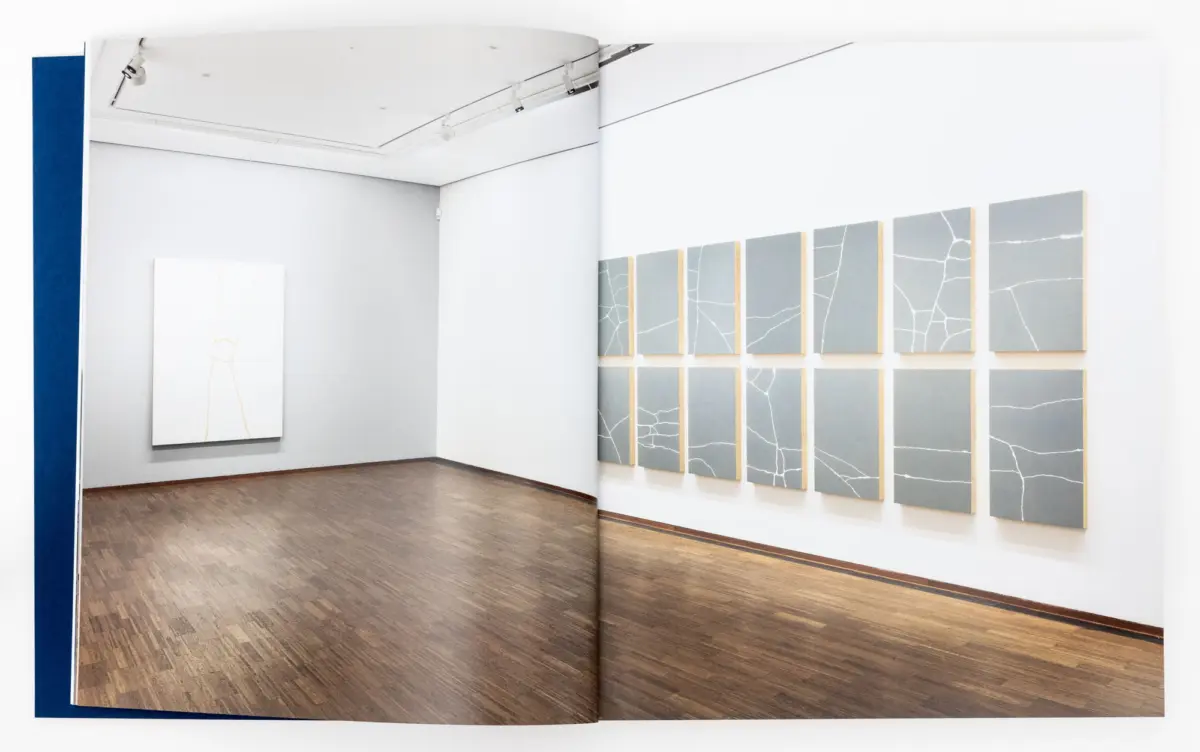 Art gallery interior with a grid of gray cracked-panel paintings on the right and a white canvas with orange lines on the left; wooden floor.