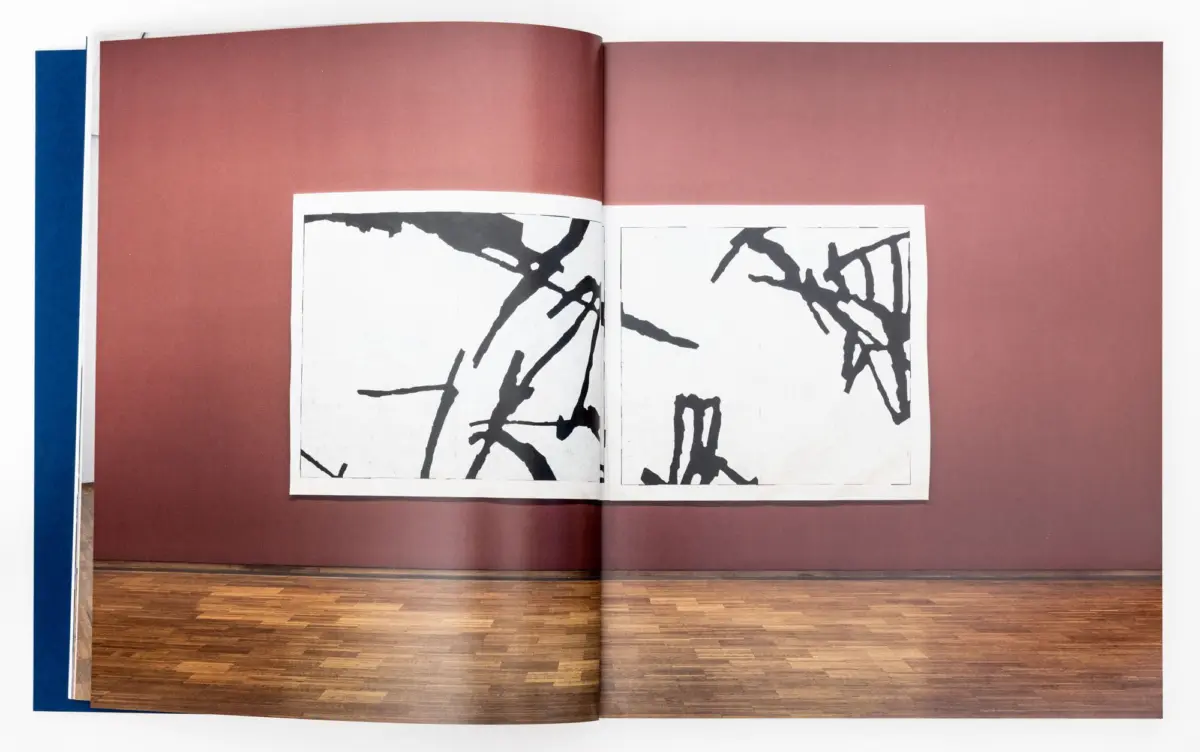 Open art book displaying two black ink abstract drawings on white pages, set against a pink backdrop with blue spine and wooden floor.