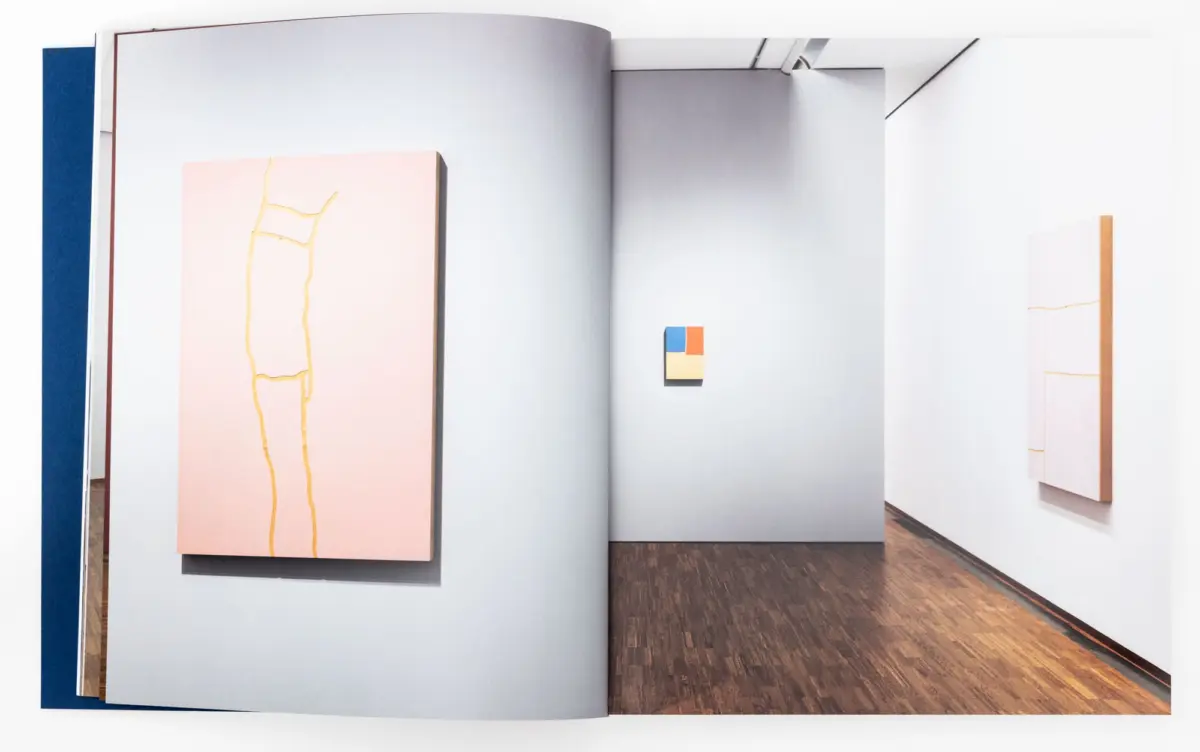 Art gallery interior: pastel pink rectangular painting with yellow lines on the left, and a small multicolor square artwork on the back wall.