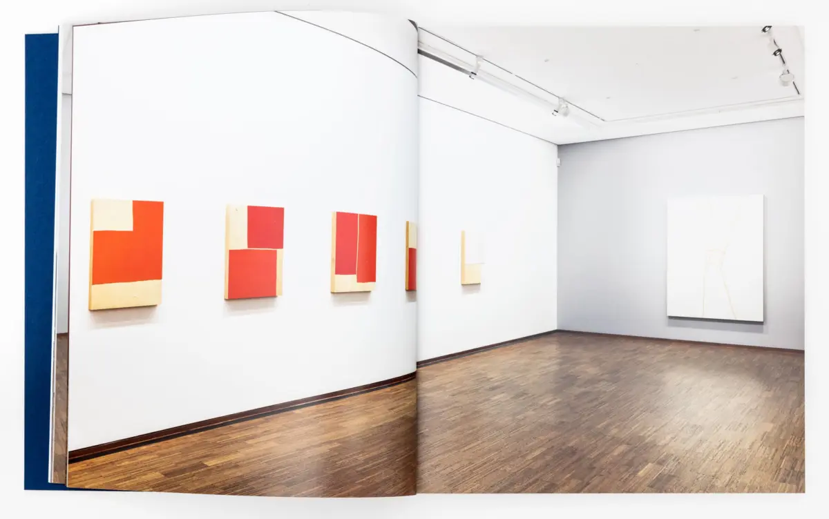 Art gallery interior with white walls and wooden floor; orange-red abstract canvases on the left and a white panel with a line drawing on the right.