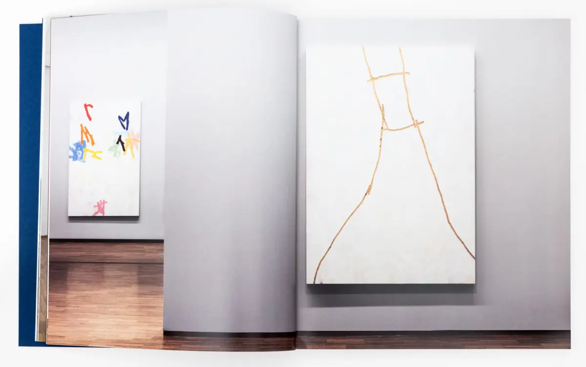 Art gallery wall with two canvases: left small colorful abstract piece; right tall white canvas with brown lines forming a figure on a wooden floor.