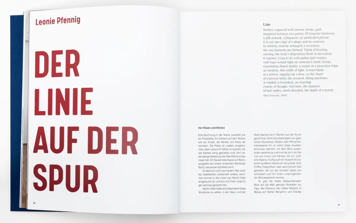 Open art book spread with bold red German headline on left page and dense text columns on right; blue spine visible.