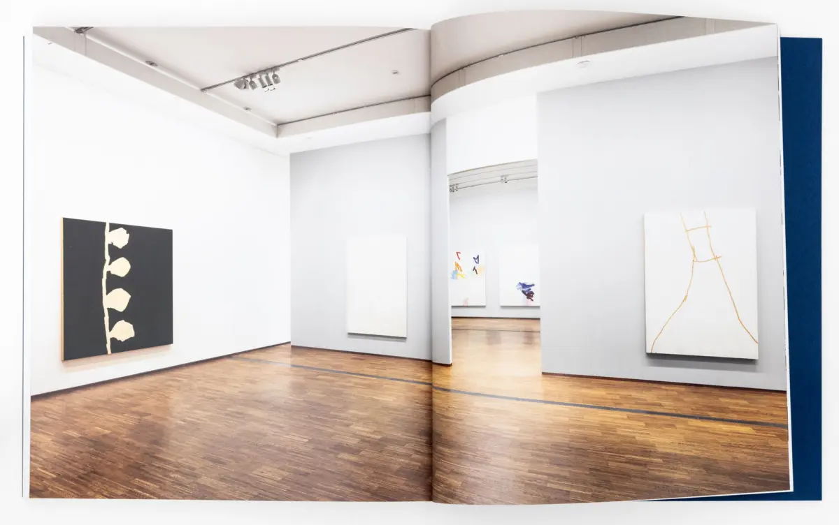 Modern art gallery interior with white walls and wooden floor, curved partitions, and abstract paintings on display.