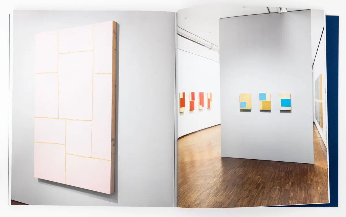 Open art catalog page showing a contemporary gallery installation: large pink panel on left, color-block artworks on grey walls.