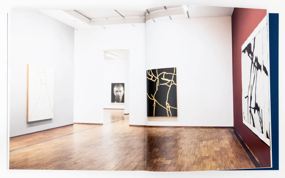 Art gallery interior with white walls and wooden floor; abstract canvases, including a black canvas with gold cracks and a white canvas with orange lines.